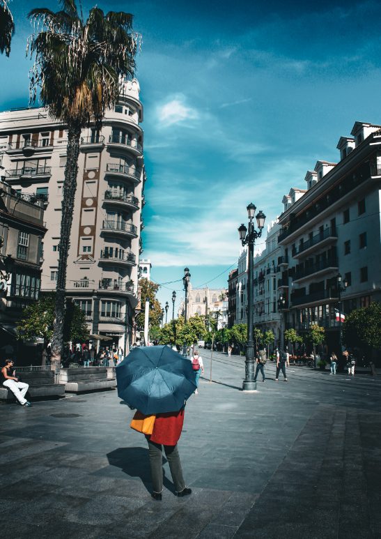 The entrance to the main street with 20th century buildings, green trees and Puerta de Jerez square where you can find very different people like someone with a colorful jacket and an umbrella.